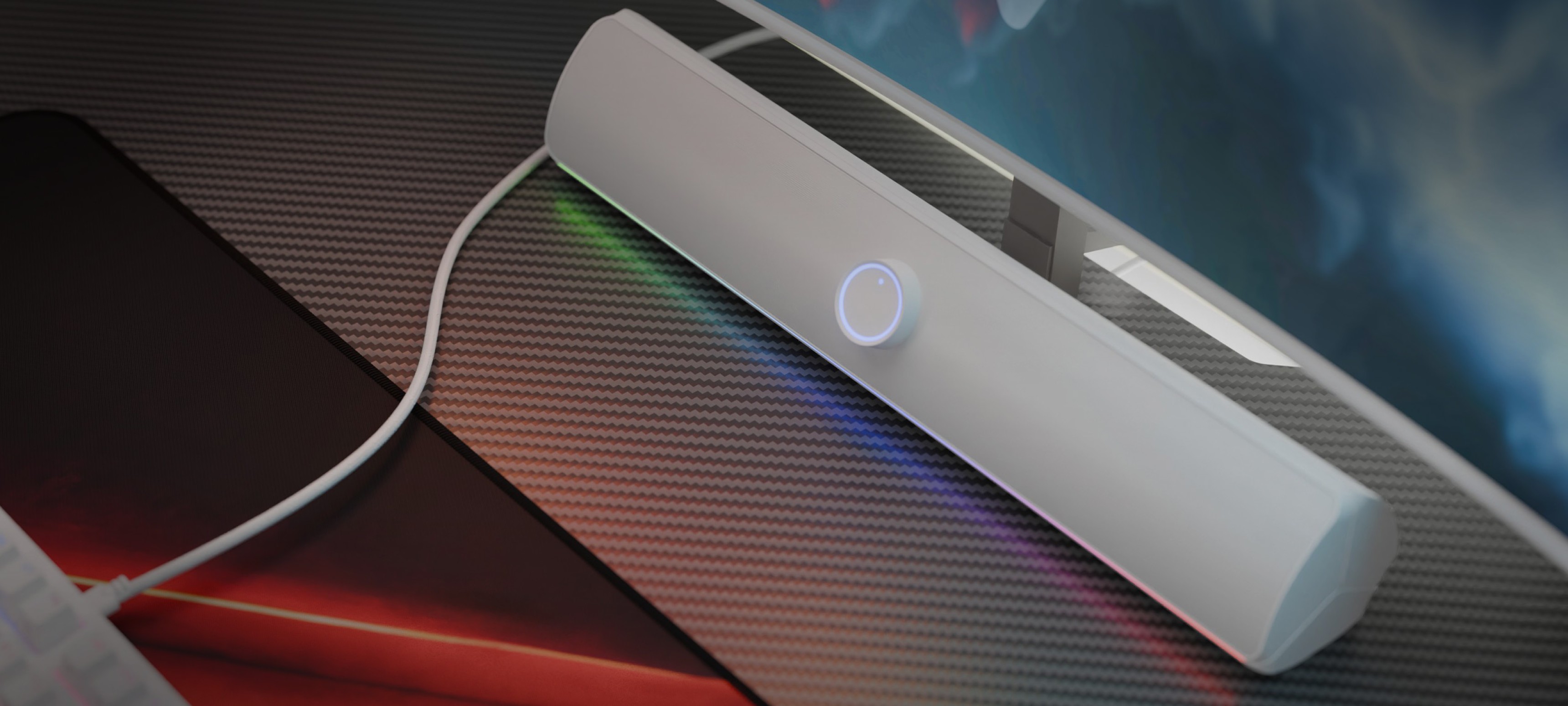 Biały soundbar Bluetooth ze świecącym przyciskiem zasilania, umieszczony na teksturowanej powierzchni obok klawiatury i monitora.
