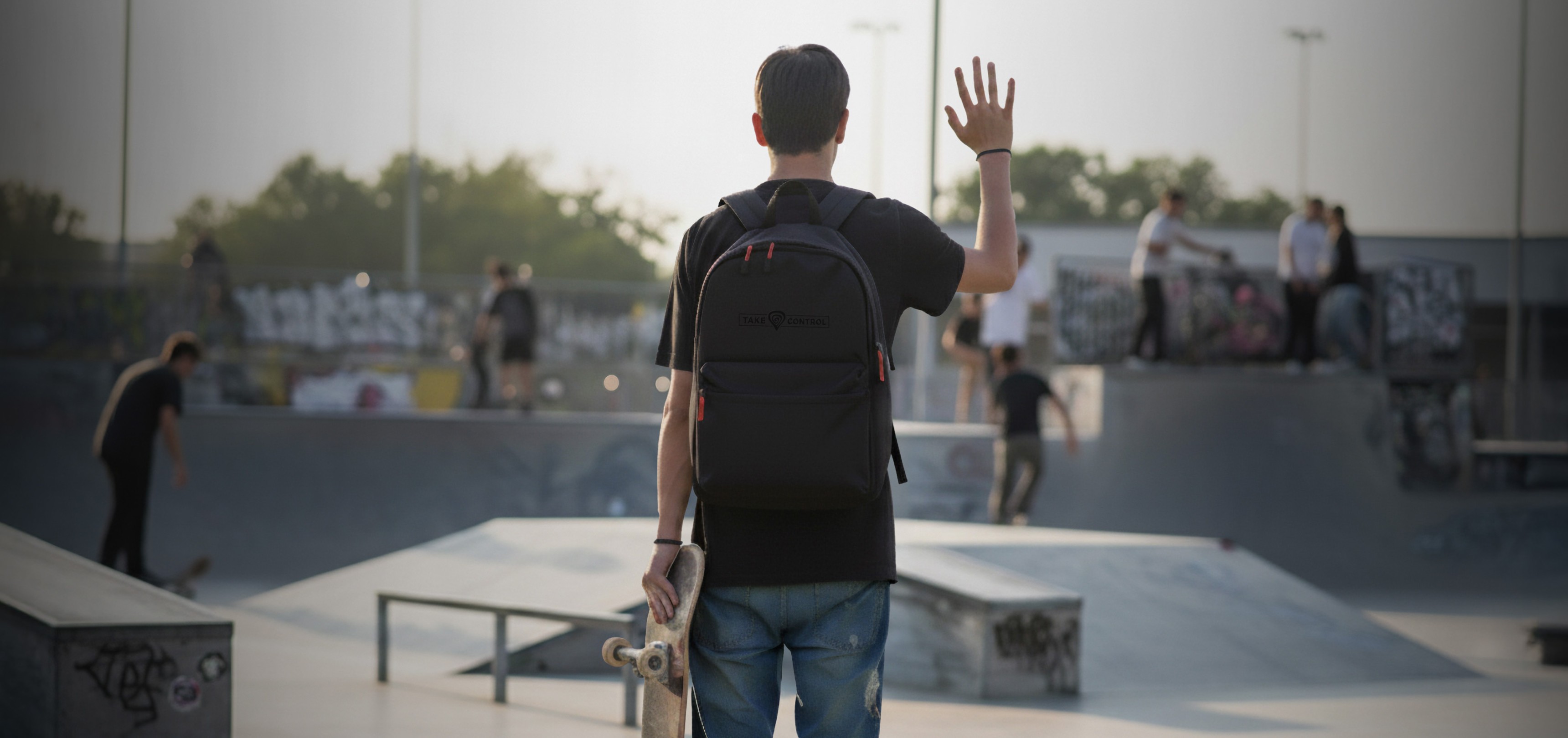 Młoda osoba w skateparku, ubrana w czarny plecak, macha trzymając deskorolkę. W tle widać skaterów i graffiti